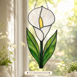 May include: A stained glass suncatcher shaped like a white calla lily flower with a yellow spadix and two green leaves. The piece is outlined in black metal cames and hangs from a thin chain. A small rectangular label reads FREE HUMMINGBIRD BONUS.