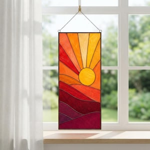 Puede incluir: Un atrapasueños rectangular de vidrieras con una escena de amanecer, un sol amarillo, rayos naranjas y rojos, y colinas superpuestas en tonos rojo oscuro y morado. La pieza está enmarcada en metal y cuelga de una cadena de tono plateado.