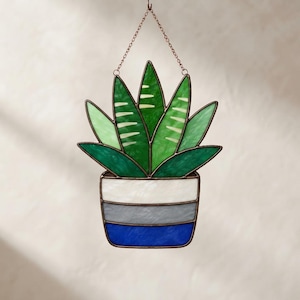 Può includere: Un acchiappasole in vetro colorato a forma di pianta succulenta in vaso. La pianta ha foglie verdi appuntite con strisce verde chiaro. Il vaso presenta tre fasce orizzontali di vetro bianco, grigio e blu, unite da profili in rame.