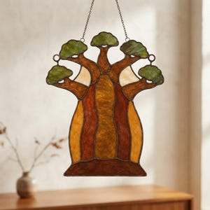 May include: A stained glass suncatcher shaped like a baobab tree, featuring a thick, textured trunk in shades of amber, brown, and gold, with five distinct branches topped with green foliage, all held together by dark metal lead lines and a chain.