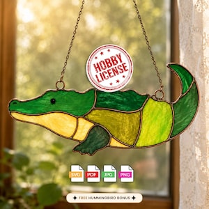 Op de afbeelding: Een alligator-zonnevanger van glas-in-lood met diverse tinten groen en geel glas, verbonden door donkere metalen soldeerlijnen. Het object hangt aan twee dunne metalen kettingen en bevat een ronde stempel met de tekst HOBBY LICENSE.