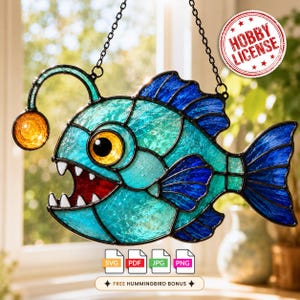 May include: A stained glass anglerfish suncatcher featuring teal and blue textured glass, a bright yellow eye, a glowing orange lure, and a red mouth with white teeth. The piece hangs from a black metal chain and includes a circular Hobby License badge.