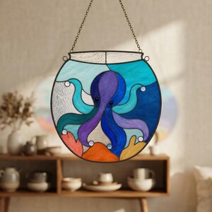 Peut inclure: Un capteur de soleil circulaire en vitrail représentant une pieuvre violette au centre d'un bocal à poissons. L'objet présente des nuances de verre bleu, orange et blanc, encadré de métal sombre avec une chaîne de suspension sur le dessus.