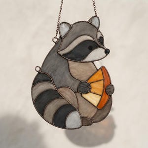 Könnte beinhalten: Ein Buntglas-Suncatcher in Form eines Waschbären mit strukturierten Glasstücken in Grau, Schwarz und Weiß. Der Waschbär hält ein dreieckiges Stück einer orangefarbenen Frucht aus durchscheinendem bernsteinfarbenem und gelbem Glas, aufgehängt an einer Kette.