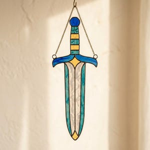 Könnte beinhalten: Ein Buntglas-Sonnenfänger in Dolchform mit einer weißen Klinge, türkisfarbenen Rändern, einem gelb-türkis gestreiften Griff und einem blauen Parierstück. Das Objekt ist an einer dünnen Metallkette befestigt, die am Griff angebracht ist.