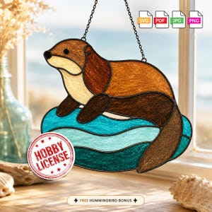 Pode incluir: Um apanhador de sol de vitral representando uma lontra castanha a descansar sobre ondas de água azul. A peça possui uma corrente de metal preto para pendurar. Um selo vermelho circular diz HOBBY LICENSE e o texto abaixo diz FREE HUMMINGBIRD BONUS.