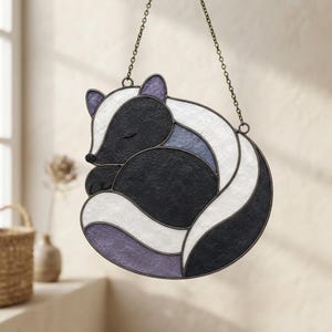 Peut inclure: Un attrape-soleil en vitrail en forme de mouffette recroquevillée, composé de pièces de verre texturées noires, blanches et violet clair assemblées par des baguettes de métal sombre. L'objet est suspendu par une fine chaîne métallique.