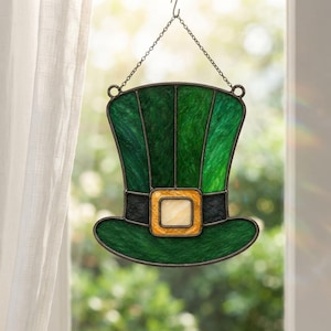 May include: A stained glass suncatcher shaped like a top hat, featuring textured dark green glass panels, a black band, and a central square buckle in a light amber hue. The piece is suspended by a thin metal chain attached to two small rings.