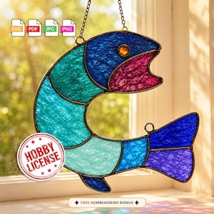May include: A stained glass fish suncatcher featuring textured blue, teal, and magenta glass panels held together by dark metal solder lines. The fish has a single amber-colored glass eye and is suspended by a thin metal chain for hanging.