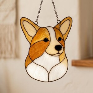 Könnte beinhalten: Ein Buntglas-Sonnenfänger in Form eines Corgi-Hundekopfes, bestehend aus bernsteinfarbenen, beigen und weißen Glassegmenten, die durch dunkle Metalllötlinien verbunden sind. Das Stück verfügt über eine Metallkette an der Oberseite zum Aufhängen.