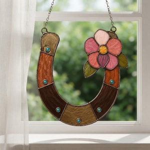 May include: A stained glass horseshoe suncatcher featuring textured panels in shades of brown, tan, and gold. The horseshoe is accented with small turquoise circular beads and a five-petaled flower in pink, purple, and yellow with green leaves.