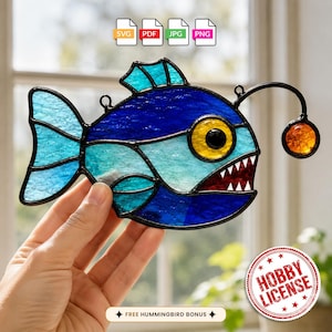May include: A stained glass anglerfish suncatcher featuring deep blue and light blue textured glass panels, a bright yellow eye, sharp white teeth, and a glowing orange lure. The design includes black metal lead lines and hanging loops.
