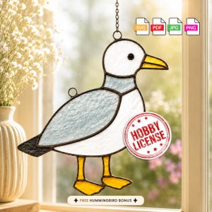 May include: A stained glass suncatcher shaped like a seagull with white, grey, and yellow glass panels held by dark metal solder lines. The design includes a red circular stamp reading HOBBY LICENSE and a label stating FREE HUMMINGBIRD BONUS.