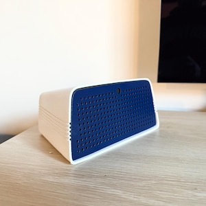 Puede incluir: Un altavoz rectangular blanco y azul con un panel frontal azul perforado. El altavoz tiene una parte superior redondeada y está sobre una superficie de madera de color claro. El altavoz tiene un diseño moderno.