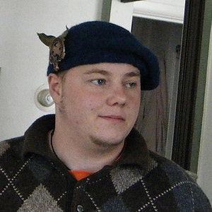 May include: A man wearing a blue beret with a decorative pin, a brown and white patterned sweater, and a light orange shirt.