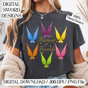 May include: A charcoal gray t-shirt featuring a graphic print of five pairs of colorful fairy wings in blue, purple, yellow, pink, and green. The text "Fairest of Friends" is printed in gold cursive in the center, surrounded by small star icons.