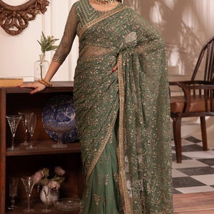 Op de afbeelding: Een olijfgroene sari met ingewikkelde gouden borduursels en een doorschijnende overlay. De sari heeft een gedetailleerde rand en is elegant gedrapeerd. Het model draagt een bijpassende blouse met doorschijnende mouwen en een gouden ketting. De achtergrond omvat een houten kast en een decoratieve spiegel.