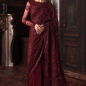 Op de afbeelding: Een bordeauxrode sari met ingewikkelde pailletten- en kanten details. De top met lange mouwen heeft doorschijnende mouwen met delicate borduursels. De sari drapeert elegant en creëert een vloeiend silhouet. De achtergrond omvat een donkere houten deur en een open haard.