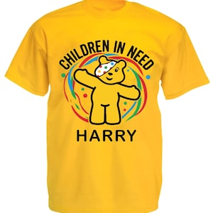 May include: Bright yellow t-shirt with the words "CHILDREN IN NEED" and "HARRY" below a cartoon bear. The bear has outstretched arms and is surrounded by a colourful design.