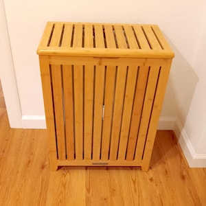 May include: A light brown bamboo laundry hamper with a slatted design. The rectangular hamper has a hinged lid with slats and vertical slats on the sides. It stands on four small feet and is placed on a hardwood floor.