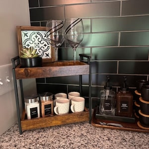 May include: A two-tiered wooden and metal serving tray with coffee and tea accessories. Includes sugar and cinnamon jars, mugs, and syrup dispensers. Two wine glasses with black accents and a decorative framed tile are also present.