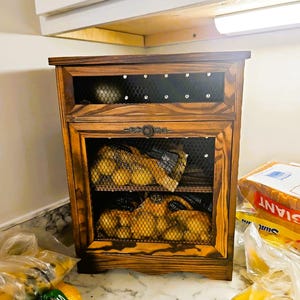 May include: A wooden kitchen cabinet with a dark stain, featuring two wire mesh compartments. The cabinet is filled with bags of potatoes. A decorative metal handle adorns the door. The cabinet is on a countertop with other produce.