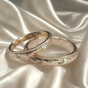Pode incluir: Duas alianças de casamento douradas com intrincadas gravuras florais. Os anéis são exibidos sobre uma superfície de cetim branco, capturando a luz e destacando os padrões detalhados. As alianças simbolizam o compromisso.