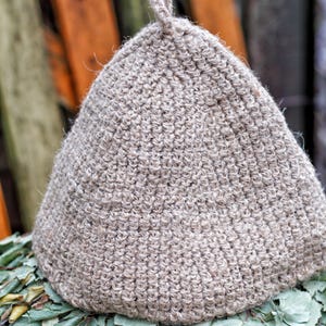 Könnte beinhalten: Ein hellbrauner, kegelförmiger Sauna-Hut aus einem strukturierten, gewebten Material. Der Hut hat eine kleine Schlaufe oben und liegt auf einem Bett aus grünen Blättern. Der Hintergrund zeigt Holzbretter.