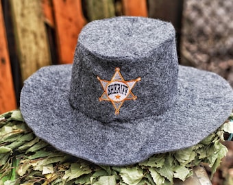 May include: A gray felt sheriff hat with a wide brim. The hat features a gold and white sheriff badge on the front. The hat is resting on a bed of green leaves.