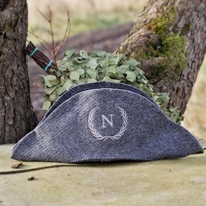 Puede incluir: Un sombrero de fieltro gris con una corona bordada blanca y la letra "N". El sombrero tiene una forma única y descansa sobre una superficie de piedra. Un manojo de ramas frondosas está en el fondo.