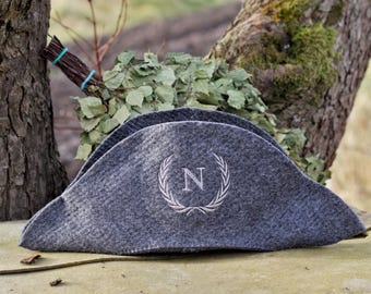 May include: A gray felt hat with a white embroidered wreath and the letter "N". The hat has a unique shape and is resting on a stone surface. A bundle of leafy branches is in the background.