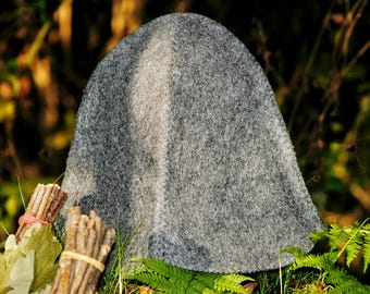 Puede incluir: Un gorro de sauna gris de lana con la parte superior redondeada, colocado entre helechos y follaje verdes. Se ven haces de ramas de abedul a la izquierda. La textura del gorro es áspera y el color es gris apagado.
