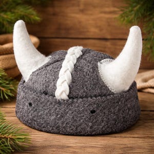 Peut inclure: Un casque de Viking en feutre gris avec des cornes blanches et un détail tressé blanc au centre. Les cornes sont incurvées et fixées sur les côtés du casque. Le casque est posé sur une surface en bois.