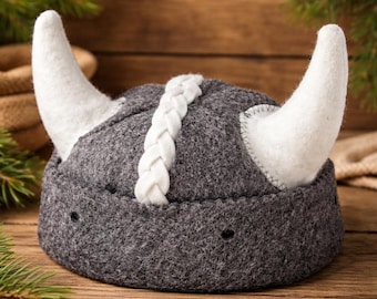 May include: A gray felt Viking helmet with white horns and a white braided detail down the center. The horns are curved and attached to the sides of the helmet. The helmet is sitting on a wooden surface.