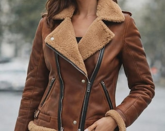 May include: A brown leather jacket featuring a tan shearling lining, a wide notched collar, and black zippered closures. The jacket includes silver-toned metal hardware, adjustable side buckles, and shearling trim at the cuffs and hemline.