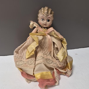 May include: Vintage doll with a painted face and molded hair. The doll wears a dress with a pink and yellow ribbon, lace trim, and a layered skirt. The doll's dress is a mix of pink, yellow, and cream colors.