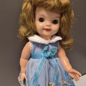 May include: A doll with shoulder-length wavy brown hair, blue eyes, and freckles. The doll wears a light blue sleeveless dress featuring a white collar, a satin ribbon waistband with two small blue fabric roses, and a mesh overlay with blue floral embroidery.