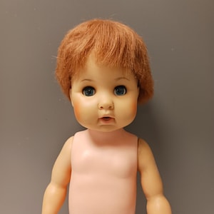 May include: A vintage baby doll with a peach-colored body and limbs. The doll has a round face with rosy cheeks, blue eyes, and a slightly open mouth. Its short, reddish-brown hair is styled in a soft, tousled manner. The doll is posed against a gray background.