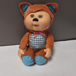 Peut inclure: Poupée vintage portant un costume d'ours brun avec des accents bleus. La poupée a un visage couleur pêche, de grands yeux et un nœud rose. Le costume d'ours a une poitrine à motifs bleus et blancs.