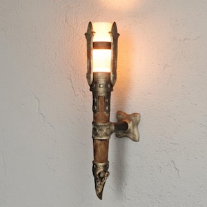 Könnte beinhalten: Eine Wandleuchte im Fackellook mit warmem, leuchtendem Licht. Die Leuchte hat einen zylindrischen Holzkörper mit Metallakzenten und einer dekorativen Halterung. Die Lichtquelle ist in einem rechteckigen, satinierten Glasschirm eingeschlossen.