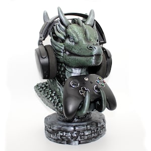 Könnte beinhalten: Eine grüne Drachenbüste mit schwarzen Kopfhörern und einem schwarzen Xbox-Controller. Die Drachenstatue hat ein detailliertes, strukturiertes Design und steht auf einem steinartigen Sockel. Die Kopfhörer sind über dem Kopf des Drachen platziert.