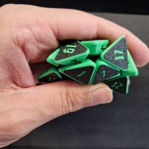 Op de afbeelding: Een verzameling groene, driehoekige dobbelstenen met zwarte, getextureerde vlakken en witte cijfers. De dobbelstenen worden in een hand gehouden, met enkele cijfers zichtbaar, waaronder 1, 12, 17 en 19. De dobbelstenen lijken 3D-geprint te zijn.