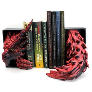 May include: Red and black dragon-themed bookends. The bookends are sculpted to resemble a dragon's head and tail, with a black base. The dragon's head has a yellow eye. Books with titles like "Monsters Know What They're Doing" are held between the bookends.