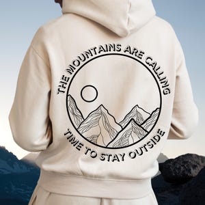Könnte beinhalten: Cremefarbener Kapuzenpullover mit schwarzem Aufdruck auf dem Rücken. Das Design zeigt einen Kreis mit Bergen, einer Sonne und dem Text "THE MOUNTAINS ARE CALLING TIME TO STAY OUTSIDE."