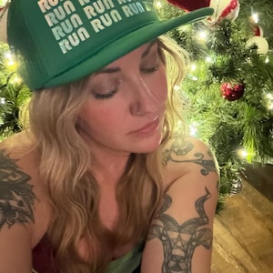 May include: A green trucker hat with the word "RUN" repeated multiple times in a white, stacked font. The hat has a mesh back and a curved brim. The hat is worn by a person with blonde hair.