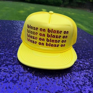 May include: A bright yellow trucker hat featuring a foam front panel and a mesh back. The front panel displays the text "blaze on blaze on blaze on blaze on" printed in a repeating pattern using a gradient of orange, red, and dark brown colors.