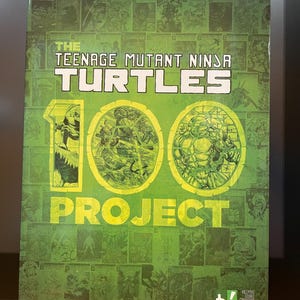 May include: A green comic book titled "The Teenage Mutant Ninja Turtles 100 Project" with the IDW logo. The cover features the title in white, two circular illustrations, and a collage of comic book panels.
