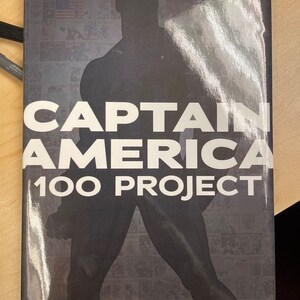 以下が含まれることがあります： コミックのパネルを背景に、キャプテン・アメリカが立っている白黒の画像。 「キャプテン・アメリカ 100 プロジェクト」というテキストが、黒地に白で印刷されている。 「ベネフィット・コミック・クリエイターズ・イン・ニード」というテキストが、画像の下部に黒地に白で印刷されている。