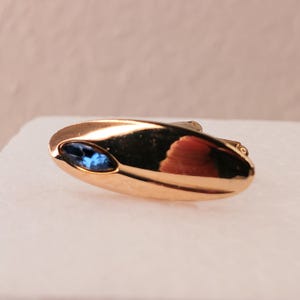 May include: A gold-toned, oval-shaped cufflink with a blue gemstone inlay. The cufflink has a smooth, polished surface and a small, circular clasp on the back. The gemstone is a vibrant blue color and is set in a bezel setting.