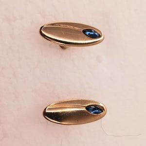 May include: A pair of oval-shaped gold-toned metal cufflinks, each featuring a smooth, polished surface with a single marquise-cut deep blue gemstone set into one end. The design includes a subtle curved line detail across the metallic face.